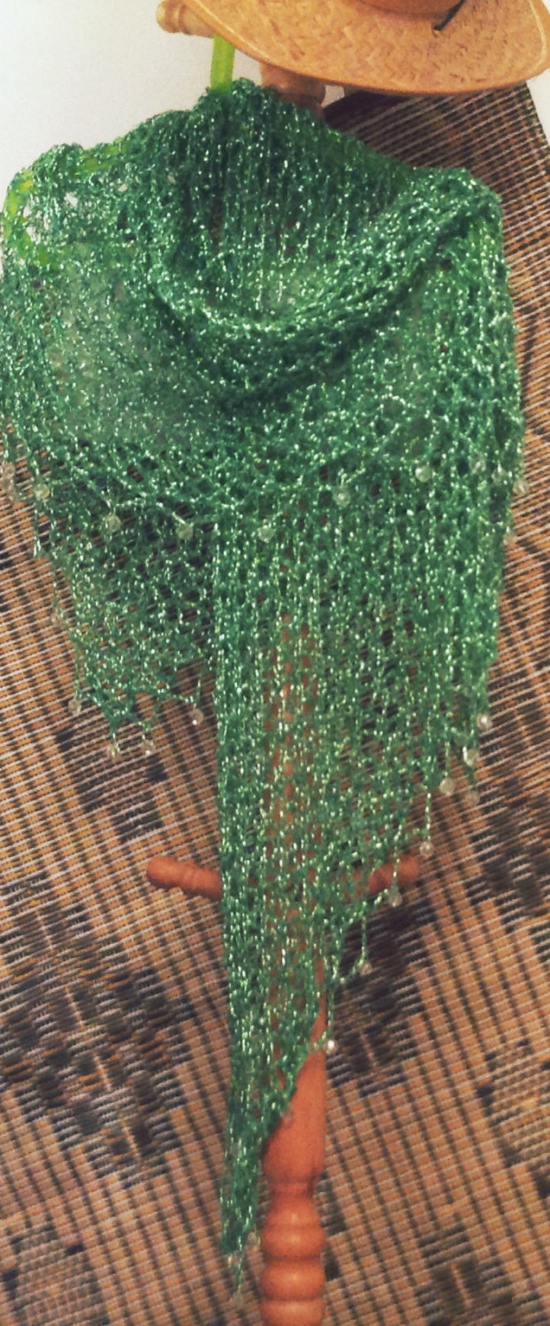 Green lurex crochet shawl hanging on a wooden stand beside a woven panel, full-length view of the shimmering wrap.