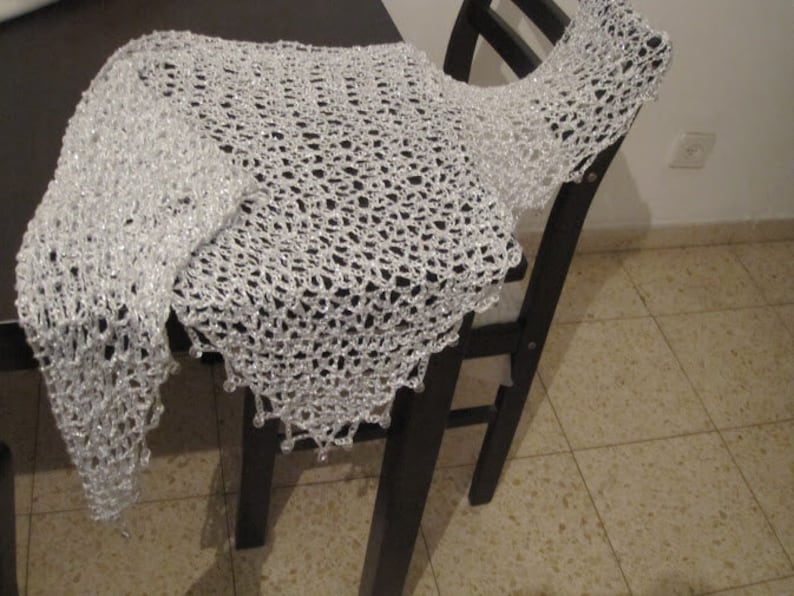 May include: A white triangular shawl featuring an openwork crochet pattern with a shimmering, metallic finish. The edges of the shawl are adorned with small, clear, dangling beads that hang from the points of the intricate, lacy knit structure.