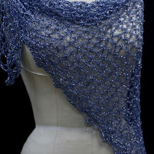May include: A periwinkle blue shawl featuring an open, loose crochet knit pattern with a subtle metallic shimmer throughout the yarn. The triangular garment drapes over the shoulders and chest, showcasing a textured, airy, and lightweight construction.