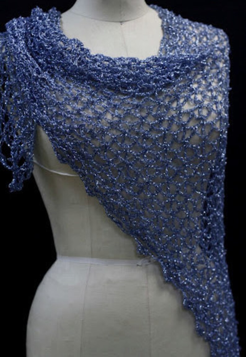 May include: A periwinkle blue shawl featuring an open, loose crochet knit pattern with a subtle metallic shimmer throughout the yarn. The triangular garment drapes over the shoulders and chest, showcasing a textured, airy, and lightweight construction.