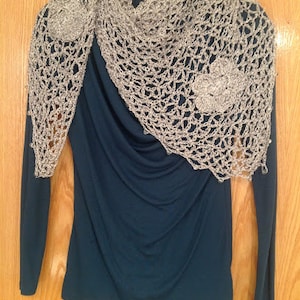 Silver lurex shawl with crocheted flower appliques draped over a navy blue top on a hanger, wooden door behind.