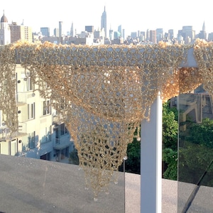 Gold lurex crochet shawl draped over a glass rooftop railing, with the New York City skyline stretching behind it in daylight.
