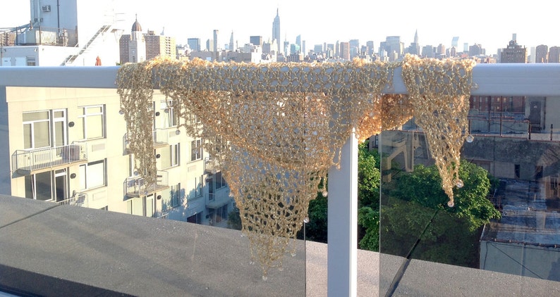 Gold lurex crochet shawl draped over a glass rooftop railing, with the New York City skyline stretching behind it in daylight.