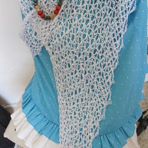May include: A white crocheted triangular shawl with an openwork pattern draped over a turquoise blue shirt with white polka dots and a white ruffled hem. A necklace featuring red, green, and tan wooden beads rests on the top of the shawl.