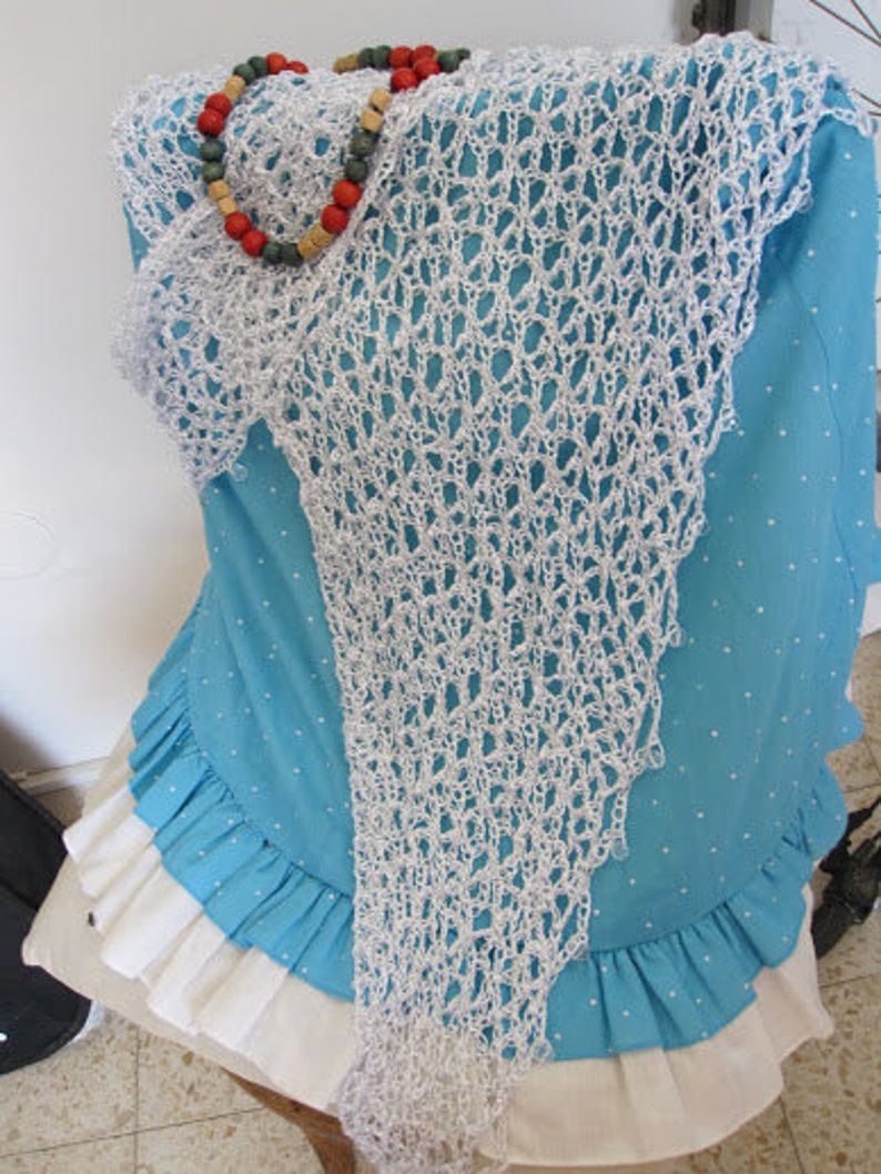 May include: A white crocheted triangular shawl with an openwork pattern draped over a turquoise blue shirt with white polka dots and a white ruffled hem. A necklace featuring red, green, and tan wooden beads rests on the top of the shawl.
