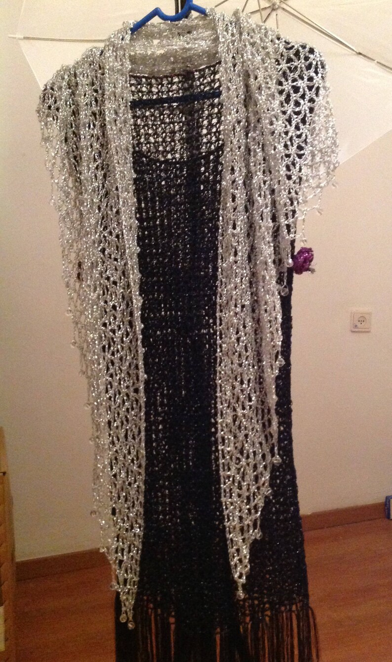 Silver lurex crochet shawl layered over a black fringed dress on a hanger, full-length view of the sparkling wrap.
