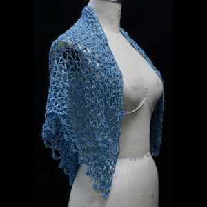 May include: A light blue crochet shawl featuring an openwork diamond pattern with a subtle metallic shimmer. The garment is draped over a beige dress form, showcasing the intricate yarn texture and the pointed, scalloped edges of the design.