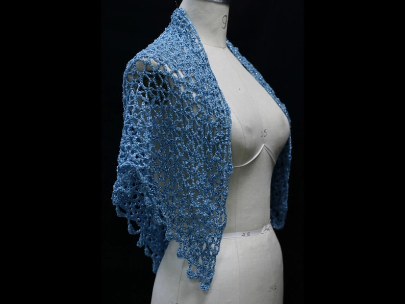 May include: A light blue crochet shawl featuring an openwork diamond pattern with a subtle metallic shimmer. The garment is draped over a beige dress form, showcasing the intricate yarn texture and the pointed, scalloped edges of the design.