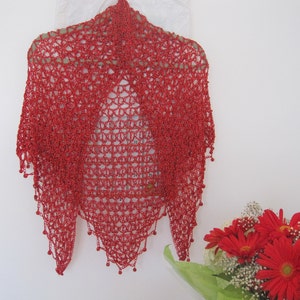 Red crochet shawl displayed flat against a white wall beside a bouquet of red flowers, showing its full triangular shape.