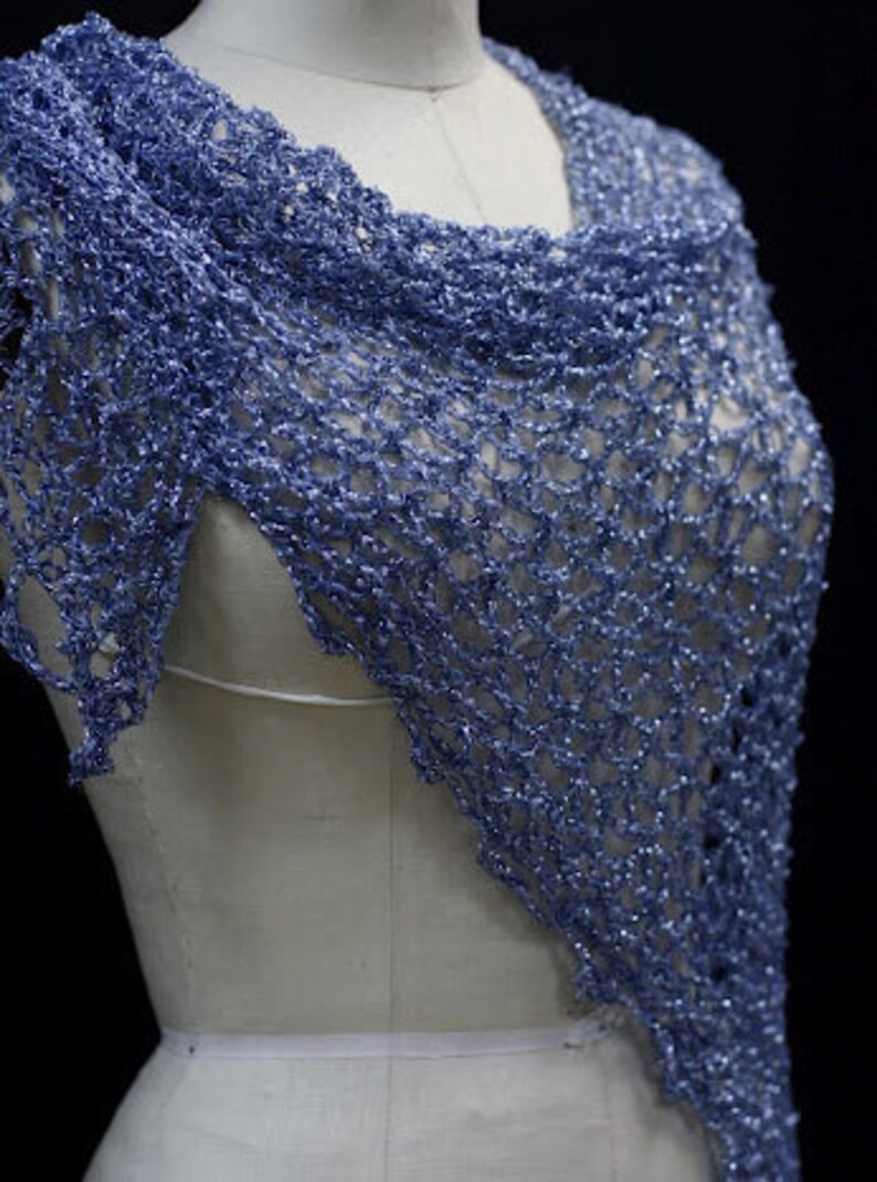 May include: A periwinkle blue shawl featuring an open, loose crochet stitch pattern with a subtle metallic shimmer throughout the yarn. The triangular garment is draped over a mannequin, showcasing the intricate, airy texture of the knit design.
