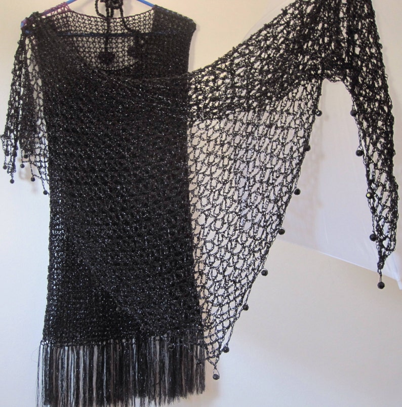 Black lurex crochet shawl draped over a black fringed dress on a hanger, side view of the shimmering wrap.