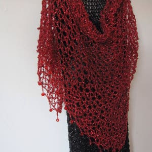 Red lurex crochet shawl draped over a black dress on a hanger, full view of the shimmering openwork wrap.