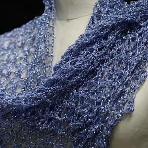 May include: A light blue scarf featuring an open, loose-knit crochet pattern with a metallic, shimmering finish. The lightweight accessory is draped over a mannequin, showcasing the intricate, airy texture of the yarn and its reflective quality.