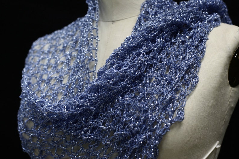 May include: A light blue scarf featuring an open, loose-knit crochet pattern with a metallic, shimmering finish. The lightweight accessory is draped over a mannequin, showcasing the intricate, airy texture of the yarn and its reflective quality.