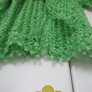 Green shawl folded on a white surface beside a flower-shaped knob, close-up of the beaded hem.