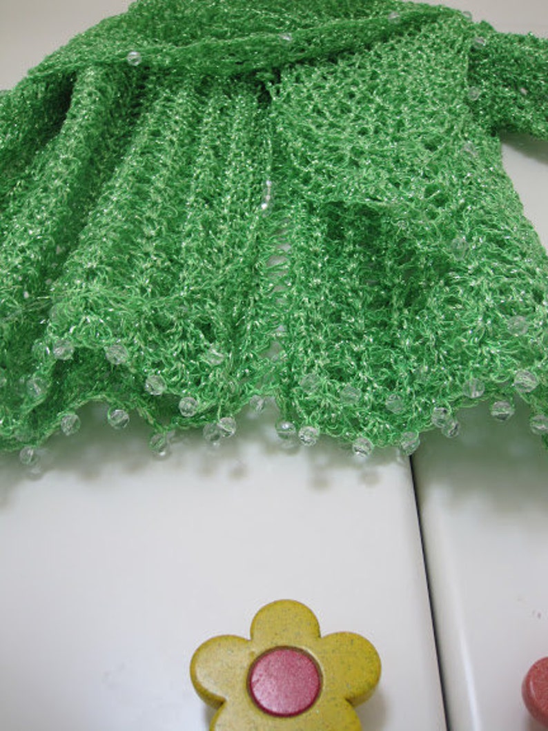 Green shawl folded on a white surface beside a flower-shaped knob, close-up of the beaded hem.