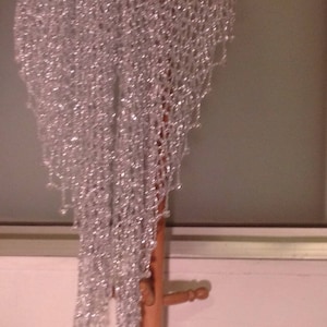Silver beaded lurex crochet shawl hanging full-length from a wooden coat stand, its shimmering openwork weave and beaded edge catching the light.