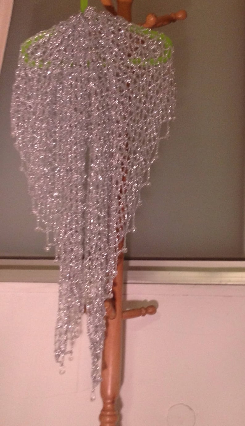 Silver beaded lurex crochet shawl hanging full-length from a wooden coat stand, its shimmering openwork weave and beaded edge catching the light.