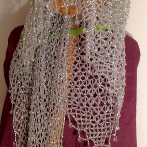 Silver lurex crochet shawl hanging on a wooden stand beneath a straw hat, long shimmering drape over a burgundy garment.