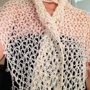 May include: A white crocheted shawl featuring an open, airy lattice stitch pattern. The edges of the shawl are adorned with small, clear, dangling beads that catch the light. The garment is draped over a solid black top and a white pearl necklace.