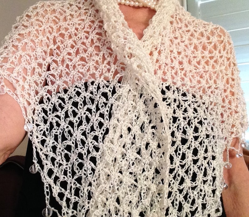May include: A white crocheted shawl featuring an open, airy lattice stitch pattern. The edges of the shawl are adorned with small, clear, dangling beads that catch the light. The garment is draped over a solid black top and a white pearl necklace.