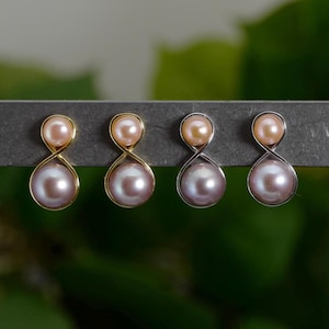 Op de afbeelding: Vier paar parel oorbellen. Twee paar hebben goudkleurige monturen en twee hebben zilverkleurige monturen. Elke oorbel heeft een kleine, perzikroze parel aan de bovenkant en een grotere, lavendelroze parel aan de onderkant, verbonden door een achtvormig ontwerp.