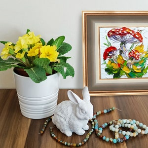 May include: A framed painting of three red and white mushrooms with green leaves and yellow flowers on a white background. A white ceramic rabbit figurine and a string of turquoise and brown beads are on a wooden table in front of the painting.