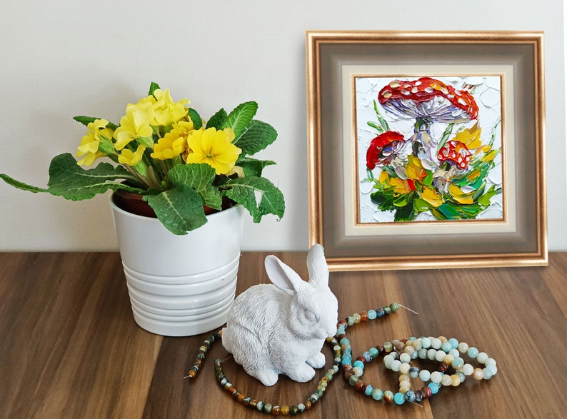 May include: A framed painting of three red and white mushrooms with green leaves and yellow flowers on a white background. A white ceramic rabbit figurine and a string of turquoise and brown beads are on a wooden table in front of the painting.