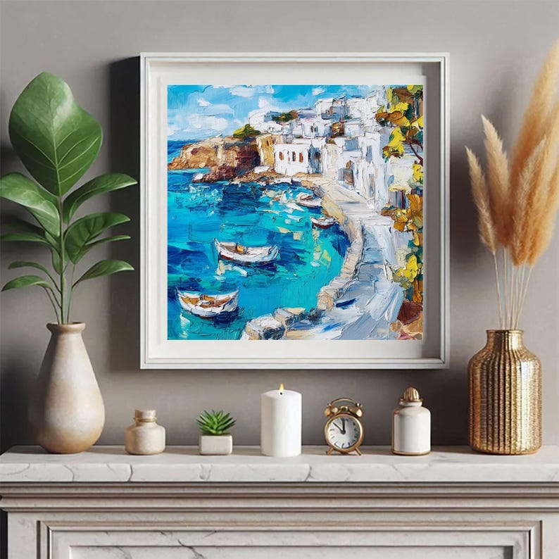 May include: A white framed painting of a seascape with white buildings on a cliff overlooking a turquoise blue ocean with white boats. The painting is on a white mantel with a white candle, a gold clock, and a gold vase with dried pampas grass.