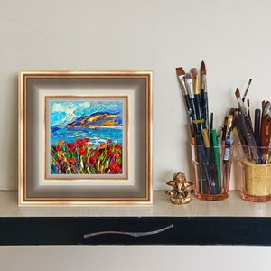 Puede incluir: Una pintura al &oacute;leo enmarcada representa un paisaje costero con amapolas rojas en primer plano, un mar azul y una cordillera bajo un cielo brillante. Pinceles en tarros de vidrio y una peque&ntilde;a estatua dorada tambi&eacute;n est&aacute;n en el estante.