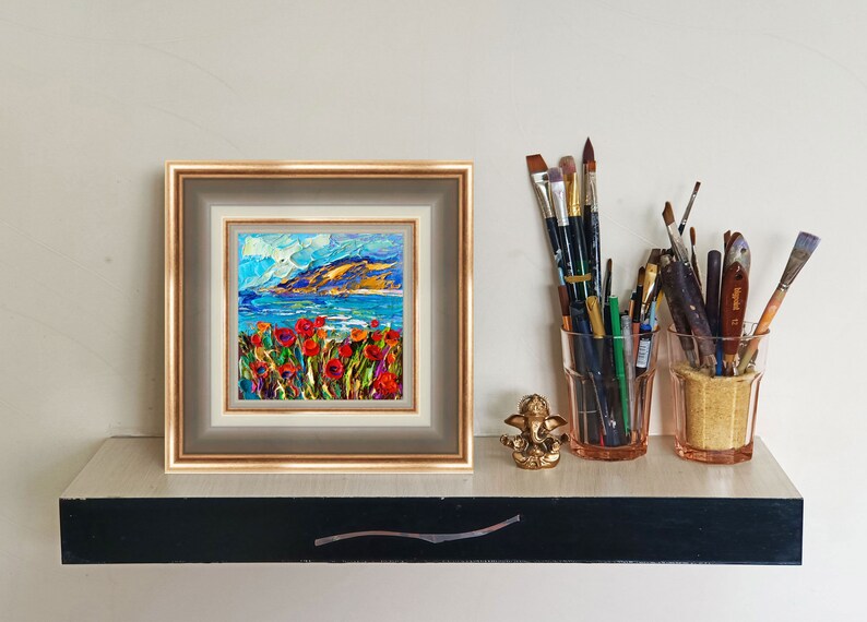 Puede incluir: Una pintura al &oacute;leo enmarcada representa un paisaje costero con amapolas rojas en primer plano, un mar azul y una cordillera bajo un cielo brillante. Pinceles en tarros de vidrio y una peque&ntilde;a estatua dorada tambi&eacute;n est&aacute;n en el estante.