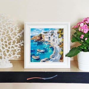 May include: A white framed painting of a coastal town with white buildings and blue water. The painting is on a shelf with a white coral sculpture and a pink flowering plant in a white pot.