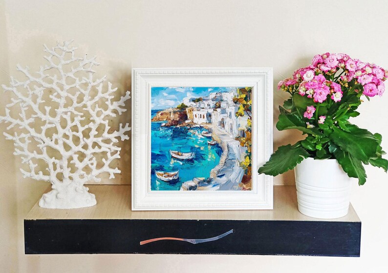 May include: A white framed painting of a coastal town with white buildings and blue water. The painting is on a shelf with a white coral sculpture and a pink flowering plant in a white pot.