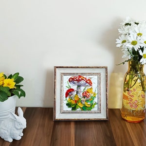 May include: A framed painting of red and white mushrooms with green leaves and yellow flowers. The frame is gold and white.