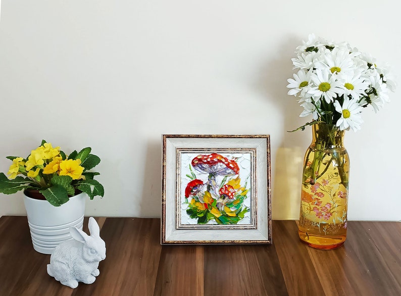 May include: A framed painting of red and white mushrooms with green leaves and yellow flowers. The frame is gold and white.