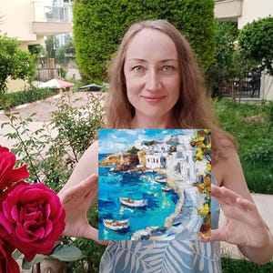 May include: A woman holds a painting of a white-washed village on a cliff overlooking a turquoise blue bay with boats. The painting is framed and has a textured brushstroke style.