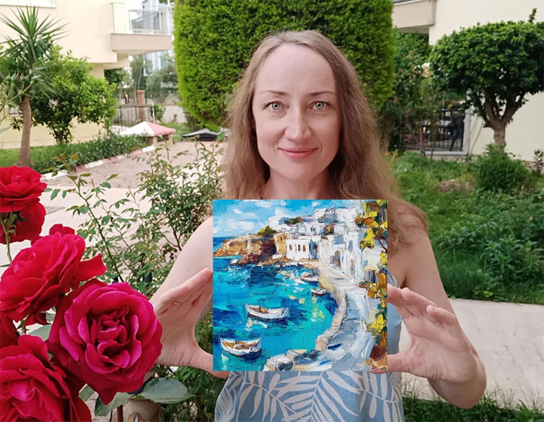 May include: A woman holds a painting of a white-washed village on a cliff overlooking a turquoise blue bay with boats. The painting is framed and has a textured brushstroke style.
