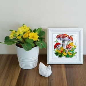 May include: A white framed painting of red and white mushrooms with green leaves on a white background. A potted yellow primrose plant is in the foreground.