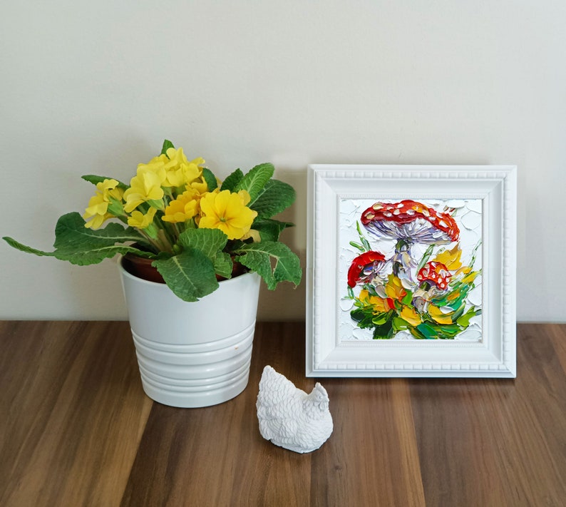 May include: A white framed painting of red and white mushrooms with green leaves on a white background. A potted yellow primrose plant is in the foreground.