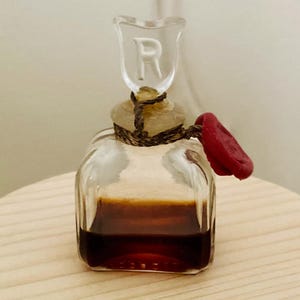 May include: A small, square, clear glass bottle containing a dark brown liquid. The bottle has a glass stopper featuring the letter R, secured with dark twine and a red wax seal attached to the neck of the bottle.