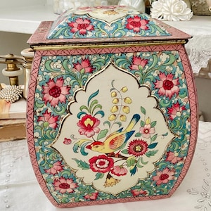 Peut inclure: Une boîte en métal vintage avec un couvercle à charnière, ornée d'un motif floral et d'oiseaux. La boîte a une bordure rose et un panneau central avec un oiseau et un motif floral en rouge, jaune et bleu. Le fond est crème.