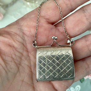 Könnte beinhalten: Eine silberne Charm-Kette mit einem kleinen Taschenanhänger. Die Tasche hat ein rautenförmiges Muster und einen Scharnierverschluss.