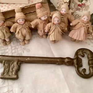 May include: A collection of antique bisque dolls wearing tan felt outfits and hats, with a large, ornate, bronze-colored key. The dolls have painted faces and are arranged on a vintage-style surface.