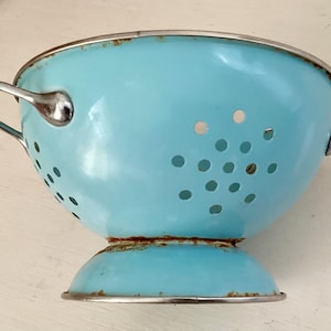 Könnte beinhalten: Eine hellblaue Vintage-Salatschüssel mit Metallrand und Griffen. Die Salatschüssel hat zahlreiche kleine Löcher zum Ablassen und ein leicht verrostetes Aussehen, was auf Alter und Gebrauch hindeutet. Die Griffe sind silberfarben.