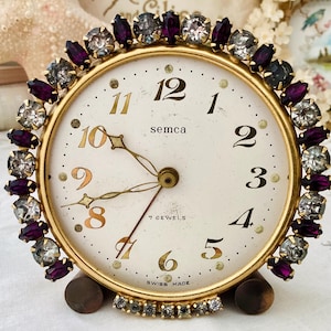 May include: A round Semca table clock with a gold-tone metal frame, featuring a white face with black numerals and gold-tone hands. The outer edge of the clock is decorated with alternating purple and clear faceted crystals in prong settings.