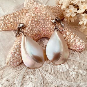 Puede incluir: Un par de pendientes de concha blanca iridiscente con cierres de clip plateados. Los pendientes tienen forma de lágrima y están colocados sobre un fondo de encaje blanco y una estrella de mar.