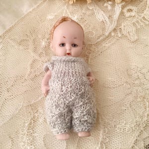 以下が含まれることがあります： グレーのニットのジャンプスーツを着た磁器製のドール。ドールは、茶色の髪と青い目をしたペイントされた顔をしています。