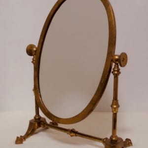 May include: A gold-toned oval tabletop mirror with a decorative stand. The mirror has a simple design with a smooth surface and a slightly ornate base.