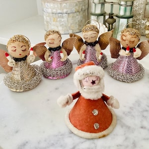May include: A collection of vintage Christmas angel ornaments and a Santa Claus figure. The angels have gold or pink skirts, gold halos, and hold candles. The Santa figure is wearing a red coat with a white fur trim.
