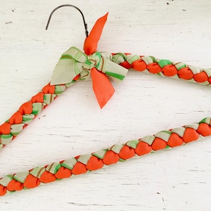 Pode incluir: Cabide decorativo de tecido trançado laranja e verde com gancho de metal. O cabide apresenta um design tecido com tecido laranja e verde e um pequeno laço no topo. Um item decorativo.
